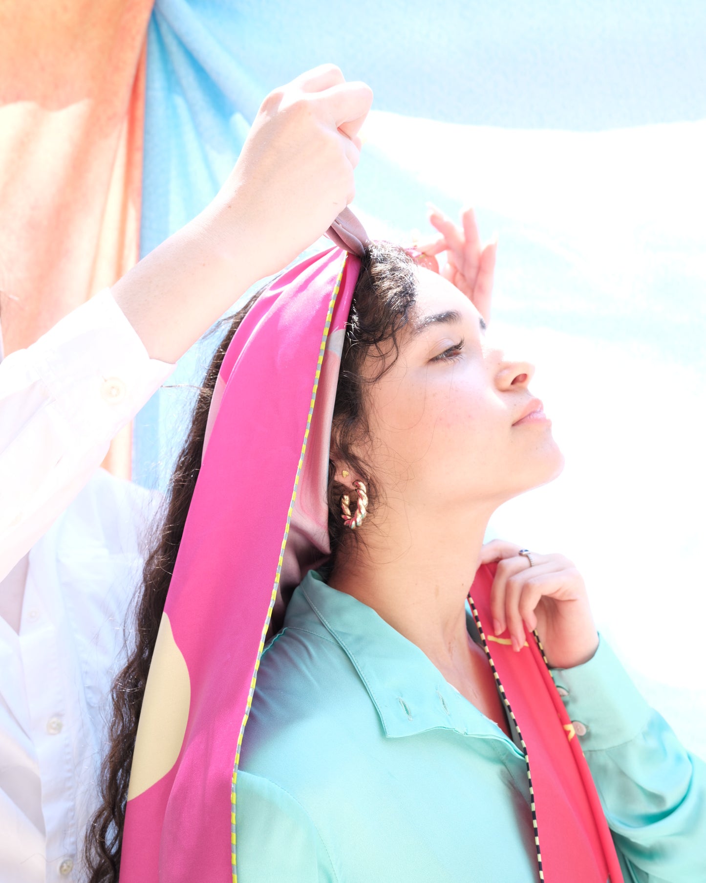 girl tying pink silk scarf on another girl outside in the sun