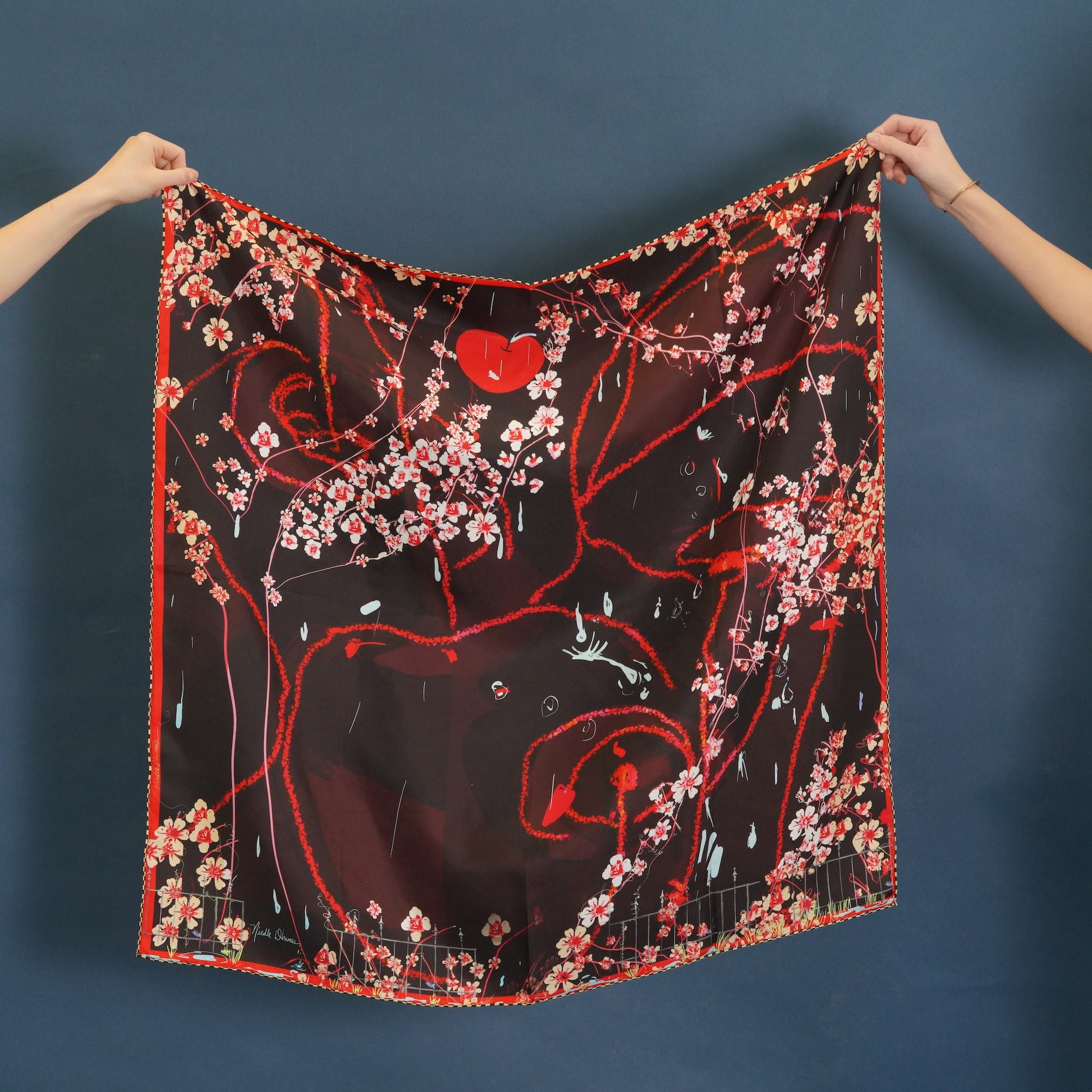 Hands holding a printed silk scarf depicting cherries and  cherry blossoms in reds, burgundy, and brown colors. 