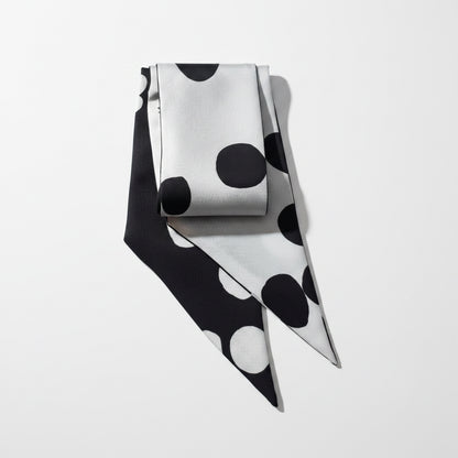A folded silk scarf with a bold black and white polka dot pattern, displayed on a textured beige linen background.