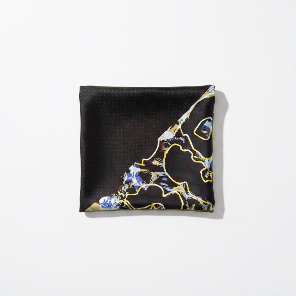 A square black scarf with a gold border, featuring a blue and gold abstract marble-like pattern in the lower right corner, laid flat on a beige fabric surface.