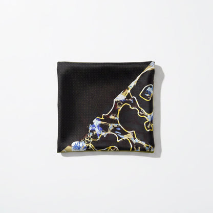 A square black scarf with a gold border, featuring a blue and gold abstract marble-like pattern in the lower right corner, laid flat on a beige fabric surface.