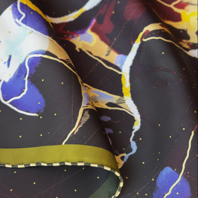 A close-up of a folded patterned fabric with a dark charcoal base, small gold dots, abstract brushstrokes in white, blue, purple, and olive green, and a dashed stitched edge.