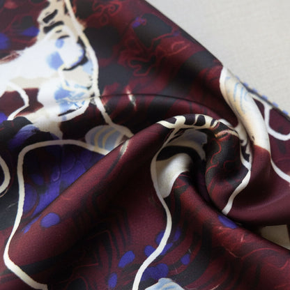 A close-up of a patterned silk scarf draped on a neutral fabric surface. The scarf features an abstract wild print design with deep burgundy, cream, black, blue, and purple elements, finished with a thin blue and white striped border.