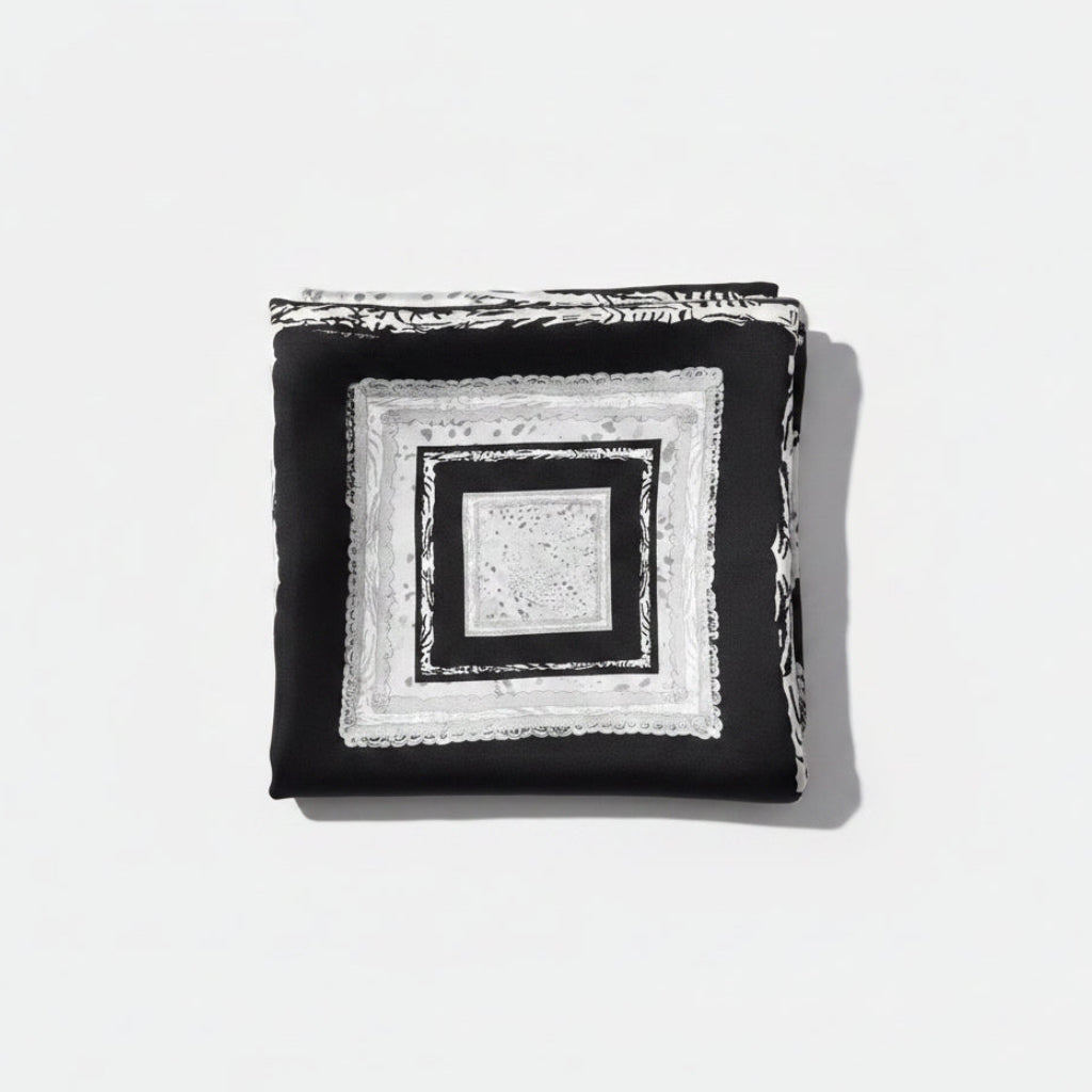 A square black and white scarf with concentric geometric and lace-like borders, laid flat on a beige textured surface.
