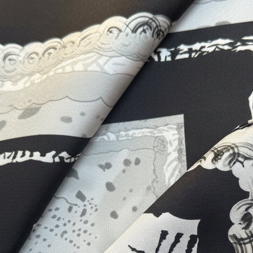 A folded square scarf with a black, white, and gray abstract pattern and ornate border details, shown on a neutral surface.