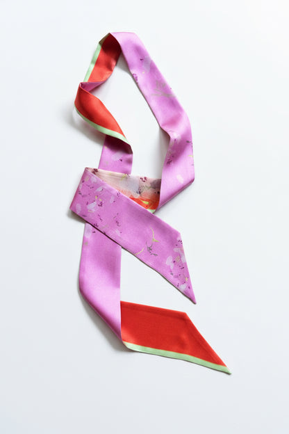 A silk scarf with a pink and orange abstract pattern and a thin light green border, styled in a loose knot on a neutral beige textured fabric background.