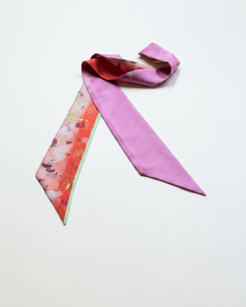 A silk scarf with a solid light pink main body and a patterned reverse side featuring orange, pink, and green floral details, laid out on a textured gray fabric surface.
