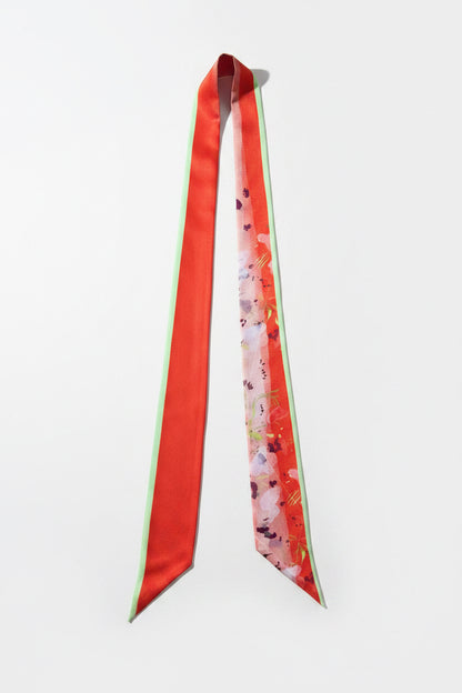 A long satin necktie laid flat on a textured gray fabric background. The tie features a solid red-orange left panel with a thin mint green edge, and a right panel with a pink and red floral pattern on a light base.