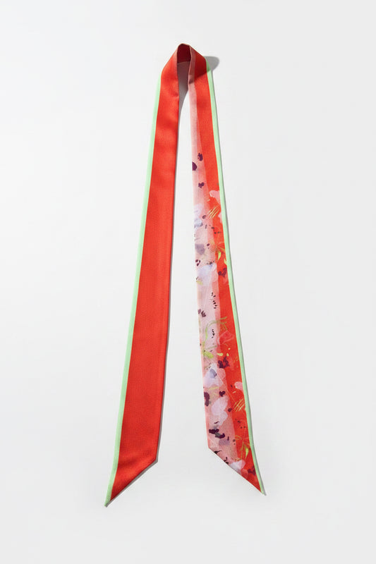 A long satin necktie laid flat on a textured gray fabric background. The tie features a solid red-orange left panel with a thin mint green edge, and a right panel with a pink and red floral pattern on a light base.