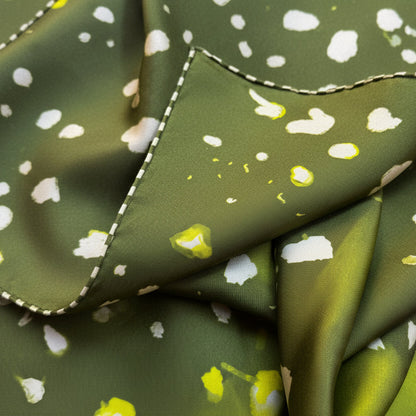 A close-up of folded olive green fabric with a white and yellow abstract spotted pattern and a visible white dashed stitched edge.