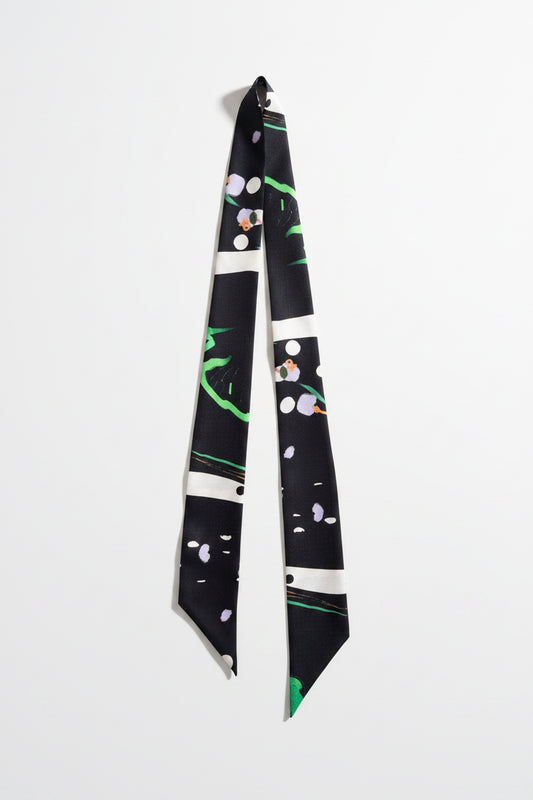 A long necktie with a black base and a modern abstract pattern featuring white stripes, green brushstrokes, and small pink and purple floral details, laid flat on a textured beige fabric background.