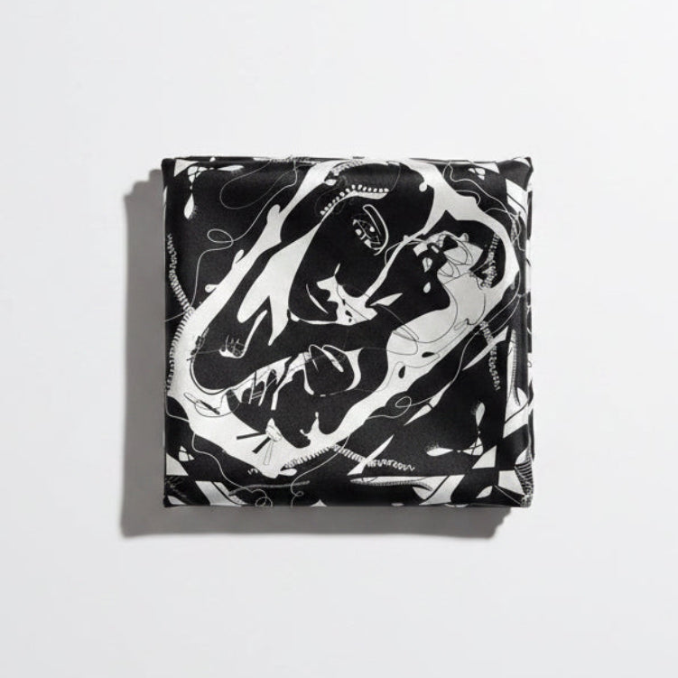 A square scarf laid flat on a beige fabric background, featuring a bold black and white graphic print with abstract faces and geometric border details.