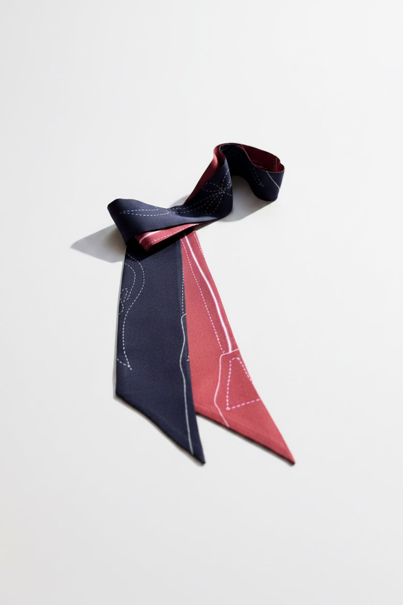 A folded fabric accessory, likely a neck scarf or tie, featuring a dark black side with white line-art sewing pattern markings and a dusty rose pink side with matching white markings, displayed on a textured light gray fabric background.